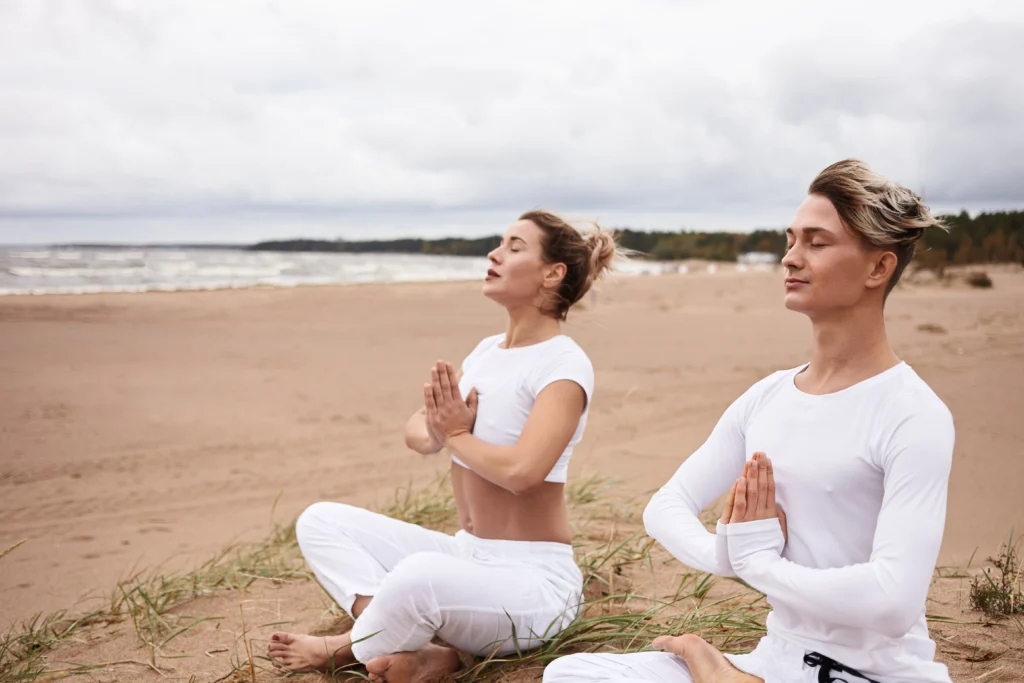 An athletic European man and woman in white sportswear sitting in Padmasana, eyes closed, holding hands in a Namaste gesture while meditating outdoors at a yoga retreat by the ocean.
