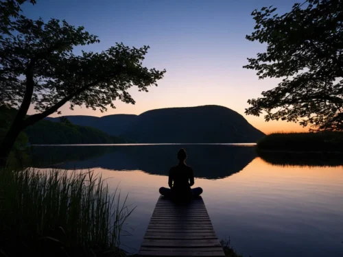 Young woman meditating by lake shore during sunset with calm water reflecting twilight sky