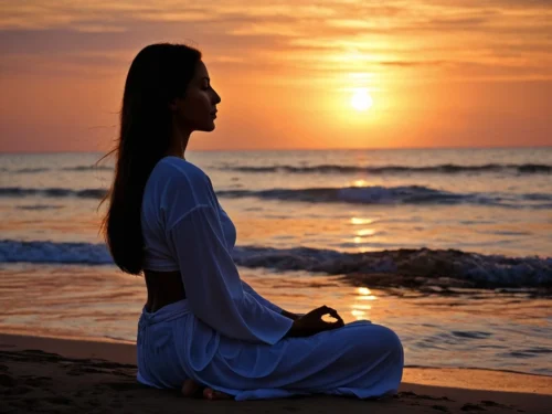 Woman in meditation pose on a beach during golden sunset, silhouetted against the warm evening sky with ocean waves in the background